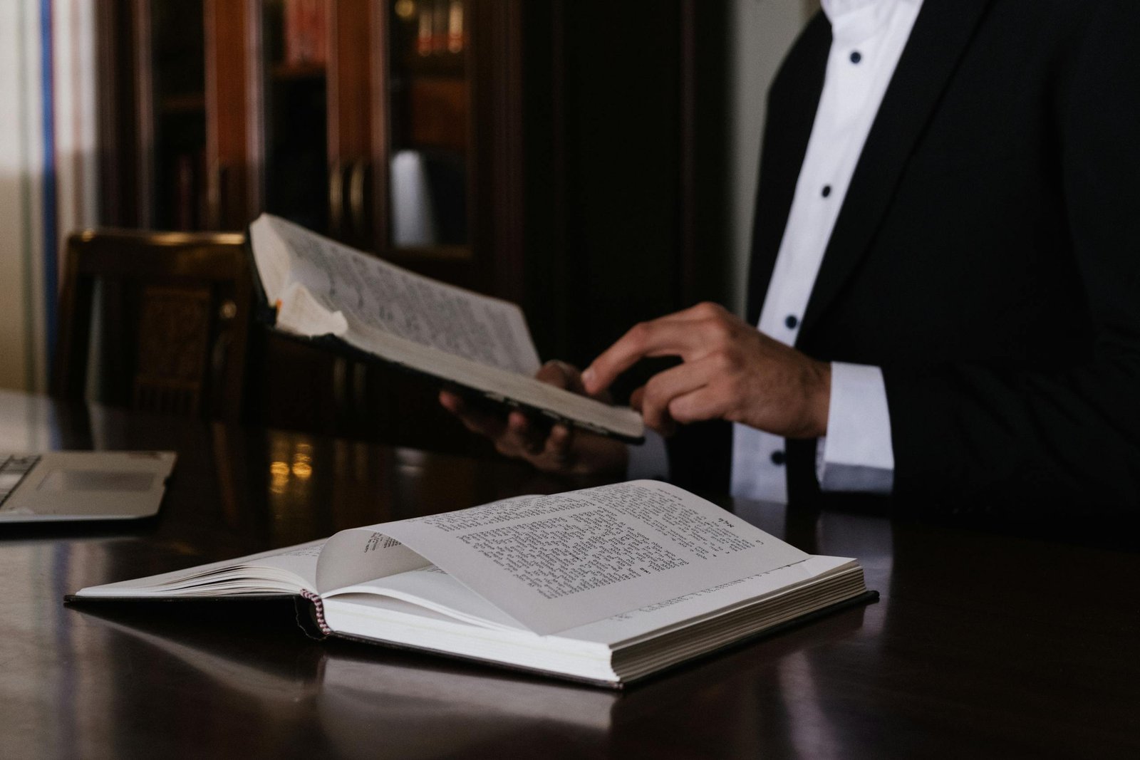 A man in a suit reading religious books at a wooden table indoors.